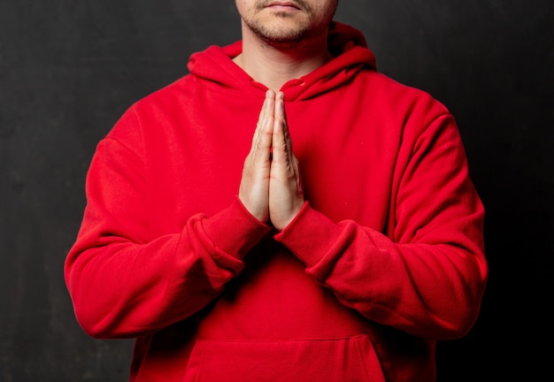 Guy en sweat-shirt rouge tenir la main en geste de prière sur un mur sombre