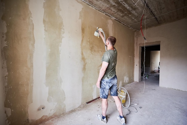 Guy tient dans la main un vaporisateur de peinture et peint un mur pendant la rénovation de la maison pendant la journée