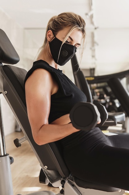 Gym nouvelle normale. Jeune femme athlétique portant un masque de prévention pendant son entraînement de remise en forme avec des haltères.