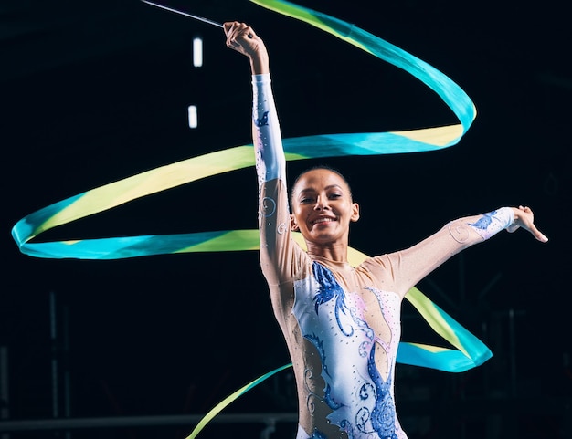 Gymnastique au ruban femme heureuse et portrait de danseuse dans un spectacle d'entraînement à la performance et une compétition dans une arène sombre Mouvement rythmique féminin et sourire pour l'action talent créatif et concert sportif