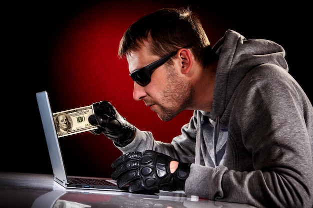 Hacker dans des lunettes de soleil avec ordinateur portable et argent en main