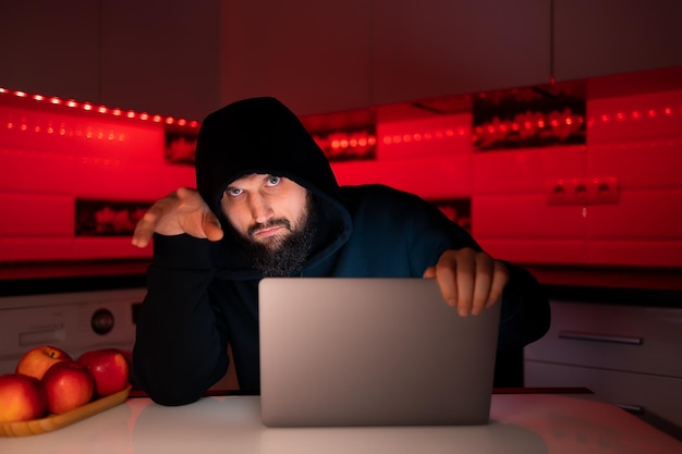 Un hacker dans un sweat à capuche noir avec une capuche sur la tête est assis devant un ordinateur portable appuyé sur son bras et regardant avec méfiance