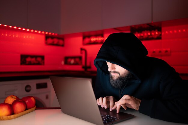 Un hacker dans un sweat à capuche noir avec une capuche sur la tête est assis devant un ordinateur portable penché sur le piratage d'Internet