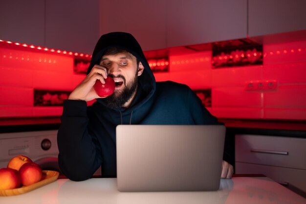 Un hacker dans un sweat à capuche noir avec une capuche sur la tête est assis devant un ordinateur portable en train de mordre une pomme rouge