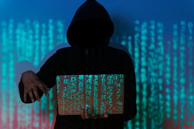 Un hacker est assis dans une pièce sombre courbé sur un ordinateur portable le hacker porte un capuchon et des lunettes de soleil