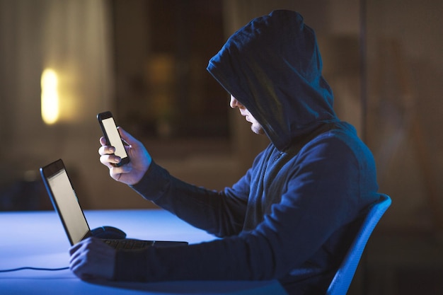 un hacker avec un ordinateur portable et un smartphone dans une pièce sombre