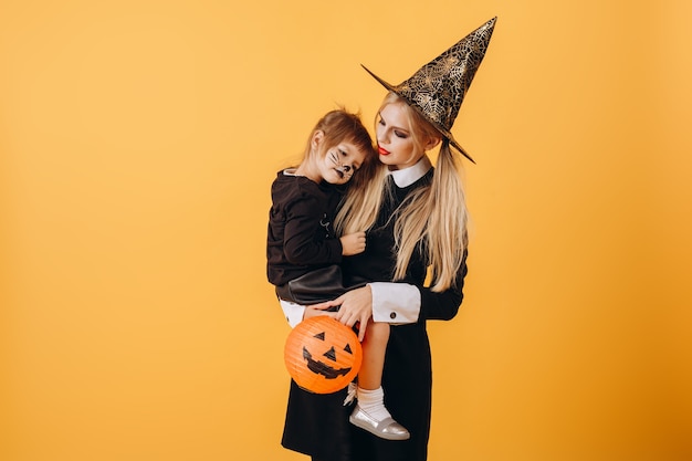 Halloween Fille Dans Un Chapeau Tient Une Petite Fille Avec Une Moustache Peinte D'un Chat Sur Un Fond D'un Mur Jaune, Copiez L'espace. Photo De Haute Qualité