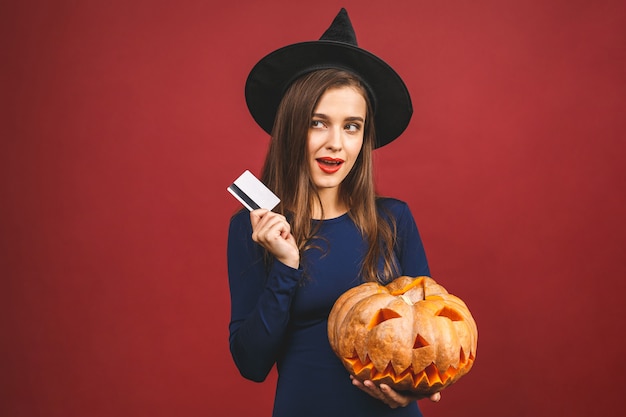 Halloween sorcière avec une citrouille sculptée et carte de crédit - isolé sur fond rouge. Émotionnelle jeune femme en costume d'Halloween.