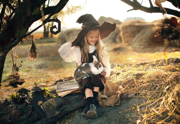 Halloween. sorcière joyeuse avec une baguette magique et un livre évoque et rit. petite fille dans un costume de sorcière.fille tenant un lapin dans ses bras