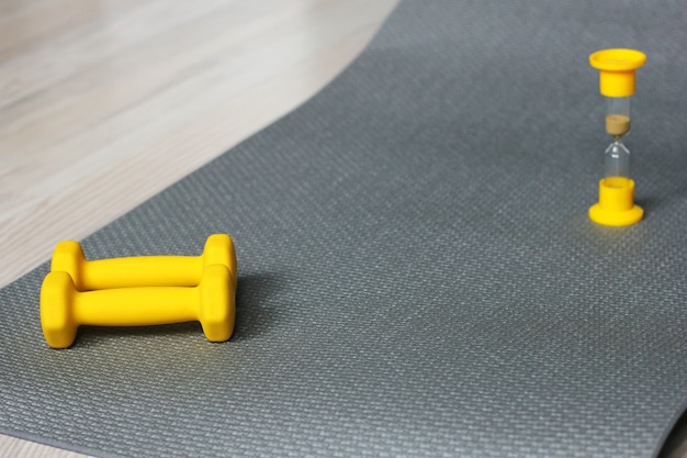 Haltères jaunes sur un tapis de fitness gris. Le sablier est à proximité. C'est l'heure du sport.