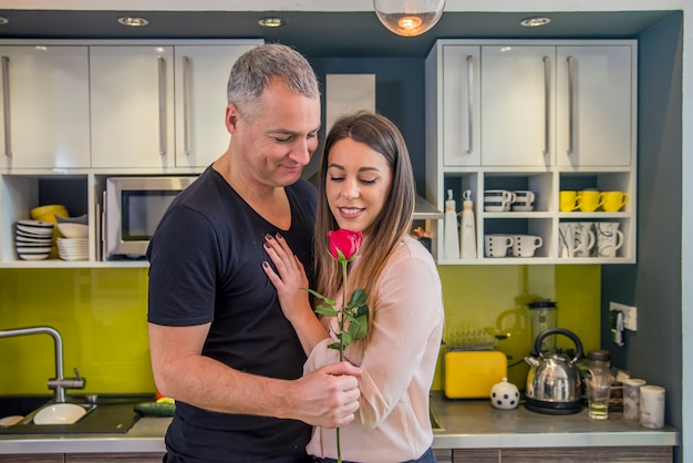 Handsome Boyfriend étreignant sa jolie petite amie avec Rose Flower et Smiling, debout dans la cuisine. Accueil romance