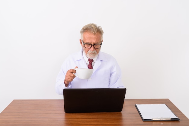 Handsome senior homme barbu médecin tenant une tasse de café tout en utilisant un ordinateur portable sur une table en bois sur blanc