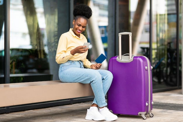 Happy Black Tourist Lady à l'aide de Smartphone assis avec valise à l'extérieur