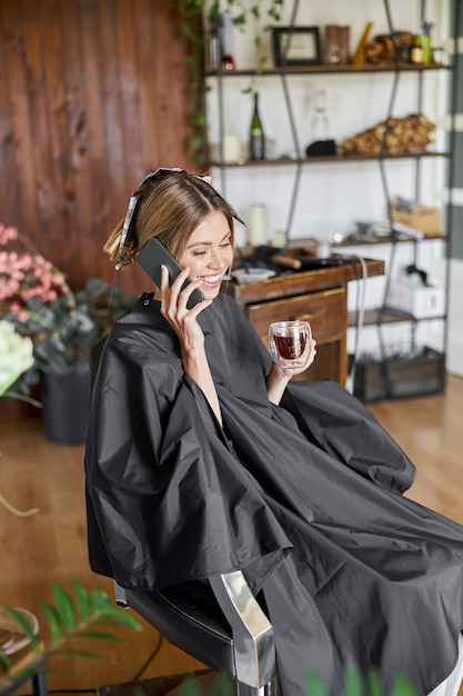 Happy blonde caucasian woman utilise un smartphone assis sur une chaise et en attente dans le salon de coiffure