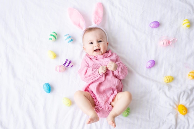 Happy Caucasian baby girl avec un bord d'oreilles de lapin sur la tête se trouve sur le dos sur le lit à la maison dans la chambre avec des oeufs de Pâques colorés bébé de Pâques