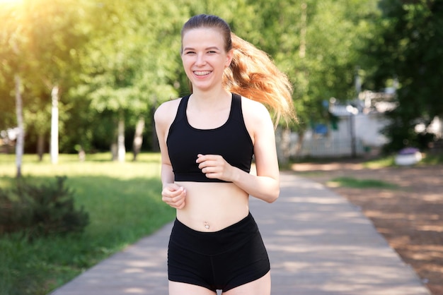 Happy fit fitness athlétique fille coureur courir jogging jeune belle femme sourit s'entraîner à l'extérieur à la journée ensoleillée d'été le matin en haut noir et chemises regardant la caméra