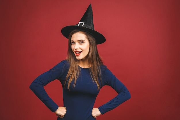 Happy Halloween Witch avec un maquillage lumineux et des cheveux longs. Belle jeune femme surprise posant en costume sexy de sorcières. Isolé sur fond rouge.