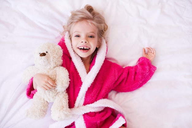 Happy kid girl holding nounours allongé dans son lit portant un peignoir