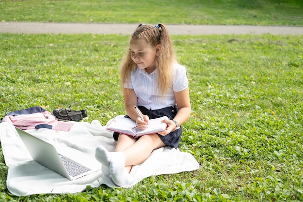 Happy kid girl on school uniform with notebook a une leçon en ligne sur un ordinateur portable sur la pelouse