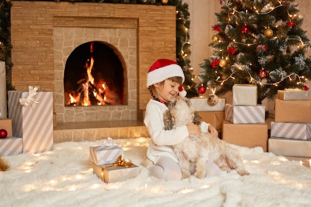 Happy kid in Christmas hat étreignant chiot, robes pull blanc et chapeau de père Noël