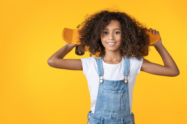 Happy preteen girl holding skateboard sur son épaule isolé sur fond jaune