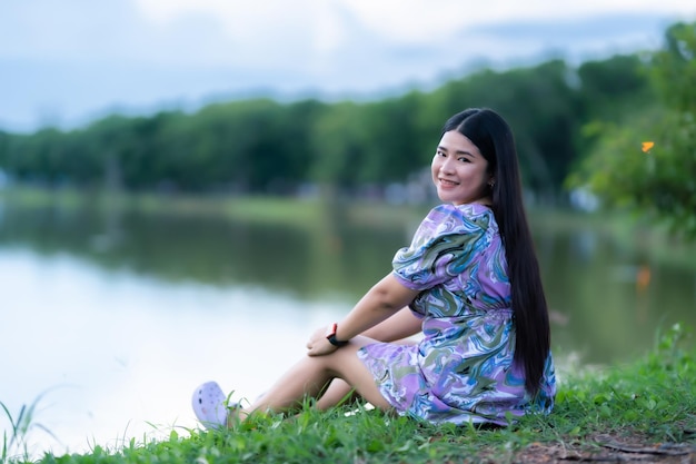 Happy Relaxing Portrait femme asiatique Porter une robe violette portant une montre intelligente tout en étant assis sur une pelouse verte à côté d'un réservoir dans le parc de la ville à l'extérieur