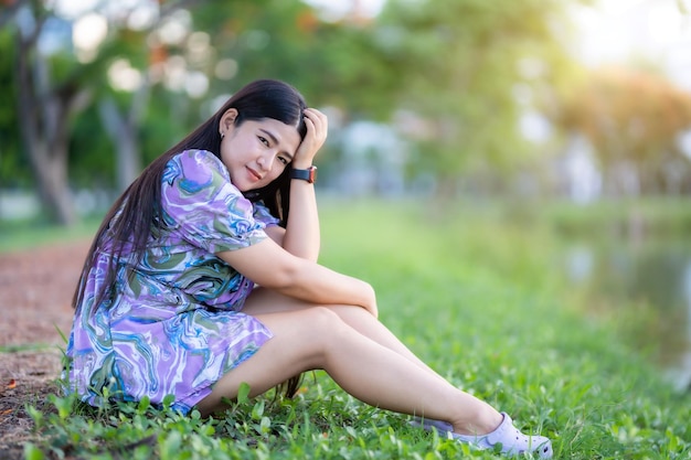 Happy Relaxing Portrait femme asiatique Porter une robe violette portant une montre intelligente tout en étant assis sur une pelouse verte à côté d'un réservoir dans le parc de la ville à l'extérieur