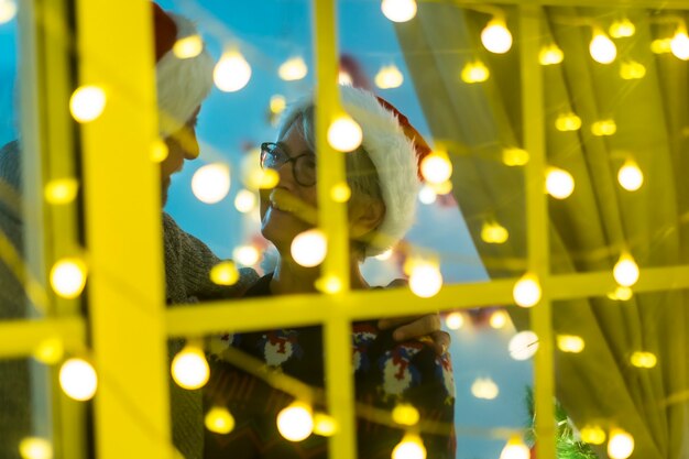 Photo happy senior couple with santa claus hats se regarde dans les yeux avec tendresse derrière la fenêtre - éclairage romantique avec de nombreuses ampoules