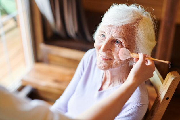 Happy senior female avec des cheveux blancs courts regardant esthéticienne pendant la procédure de maquillage dans un salon de beauté