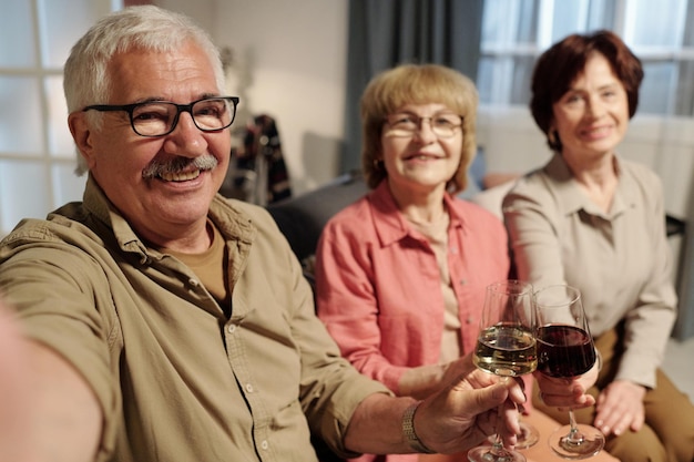 Happy senior man in eyeglasses tinter avec un verre de vin tenu par deux femmes