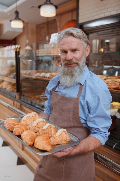 Happy senior man travaillant dans sa boulangerie, transportant un plateau avec des croissants