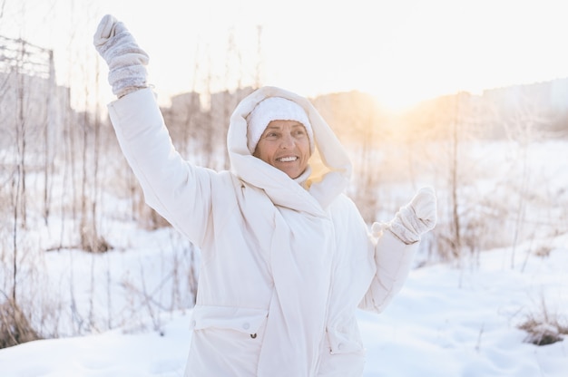 Happy senior senior woman in white outwear chaud jouant avec la neige en plein air d'hiver ensoleillé