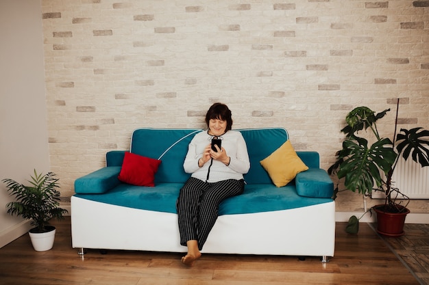 Happy senior woman sitting sur le canapé bleu utilise le téléphone pour un appel vidéo