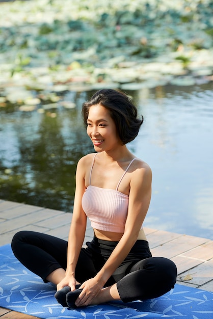 Happy slim fit jeune femme assise sur un tapis de yoga en position du lotus sur une jetée en bois