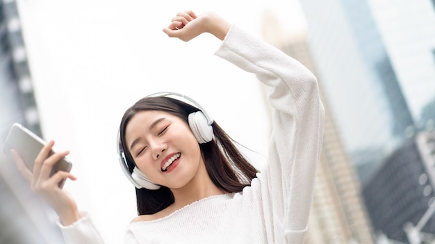 Happy smiling Asian girl wearing headphones écouter et passer à la musique contre le mur du bâtiment de la ville