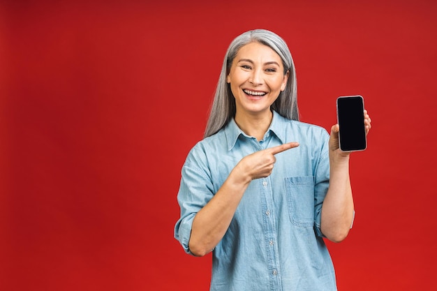 Happy smiling asian senior mature woman in casual montrant un écran de smartphone vierge tout en regardant la caméra isolée sur fond rouge à l'aide de téléphone