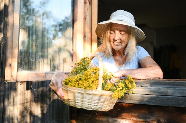 Happy smiling femme senior émotionnelle posant par fenêtre ouverte dans la vieille maison de village en bois en chapeau de paille avec panier de fleurs