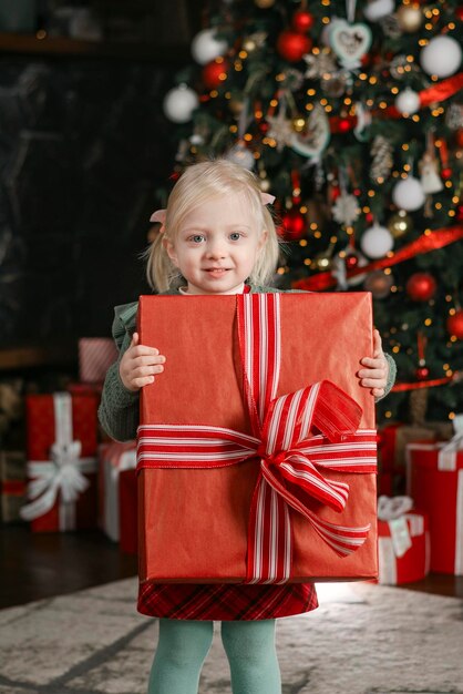 Happy threeyears old girl est titulaire d'une grande boîte-cadeau et se penche sur l'appareil photo sur fond d'arbre de Noël cadre vertical de la veille de Noël