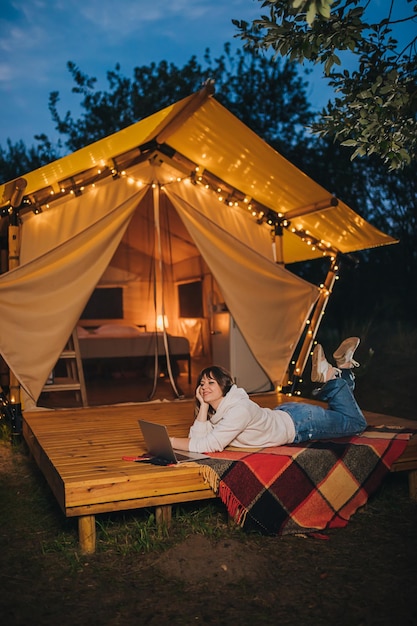 Happy Woman pigiste à l'aide d'un ordinateur portable sur une tente de glamping confortable dans une soirée d'été Tente de camping de luxe pour des vacances en plein air et des vacances Concept de style de vie