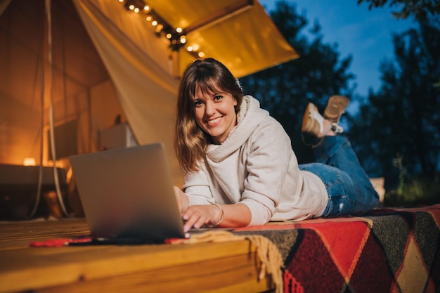 Happy Woman pigiste à l'aide d'un ordinateur portable sur une tente de glamping confortable dans une soirée d'été Tente de camping de luxe pour des vacances en plein air et des vacances Concept de style de vie