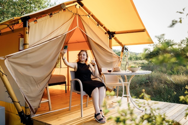 Happy Woman pigiste parlant avec des amis via un appel vidéo tout en se relaxant sur une tente de glamping confortable en journée ensoleillée Tente de camping de luxe pour les vacances d'été en plein air et les vacances Concept de style de vie