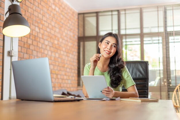 Happy young Asian businesswoman implantation dans son bureau de salon à domicile souriant tout en pensant à ses idées d'affaires