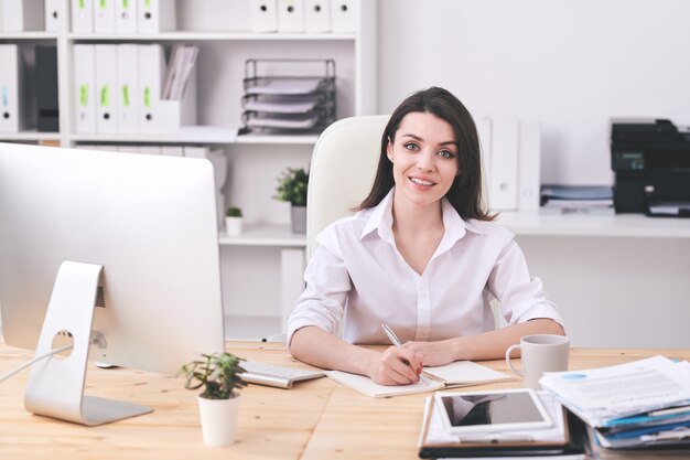 Happy young brunette businesswoman in formalwear vous regarde tout en écrivant le plan de la journée de travail le matin au bureau