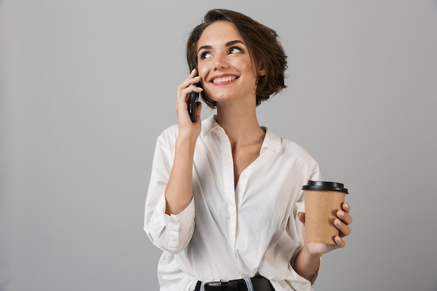 Happy young business woman posing isolé sur mur gris parler par téléphone mobile et boire du café