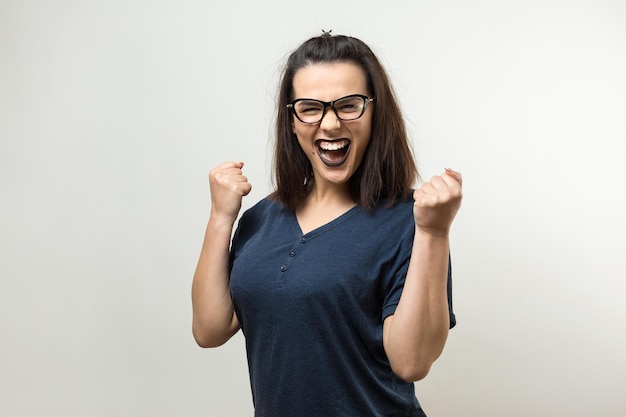 Happy young caucasian femalein dans une chemise bleue avec des lunettes lève un poing fermé pour la joie