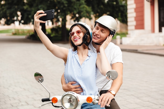 Happy young couple in crash casques faisant selfie sur smartphone alors qu'il était assis ensemble sur le scooter à l'extérieur