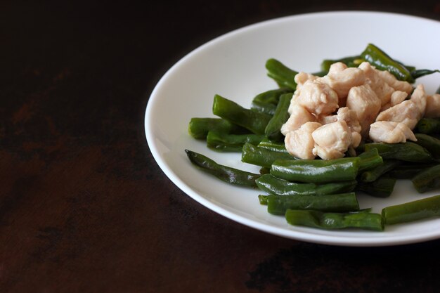 Haricots verts et morceaux de poitrine de poulet dans une assiette blanche sur un comptoir en bois.