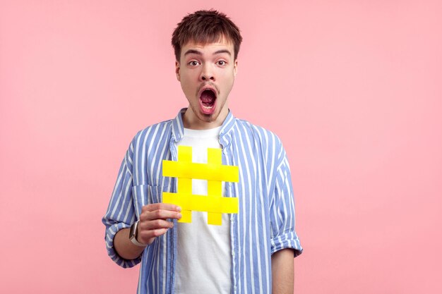 Hashtag pour les dernières nouvelles. Portrait d'un homme aux cheveux bruns choqué en chemise décontractée tenant un grand grand panneau de hachage jaune l'air surpris, wow expression. studio d'intérieur tourné isolé sur fond rose