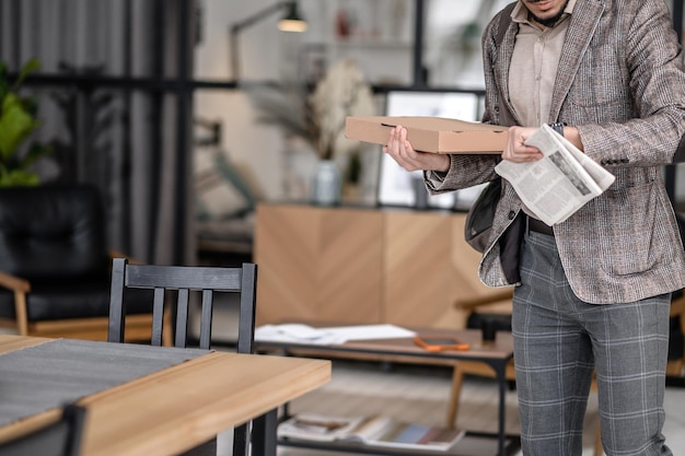 Hâtivement. Homme en costume tenant un journal et une boîte regardant une montre-bracelet debout près de la table à la maison sans visage