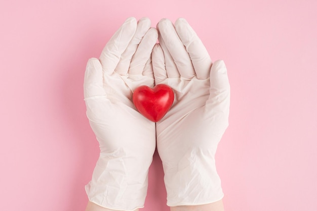 En haut au-dessus des frais généraux de la première personne vue rapprochée photo de coeur rouge dans les mains d'une femme médecin dans des gants isolés sur fond rose pastel avec fond